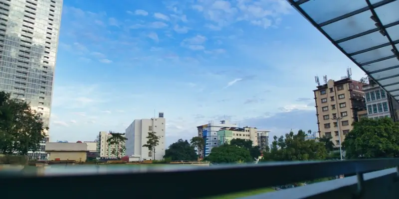 A view of several mid rise buildings and trees under a bright blue sky, seen from behind a balcony railing and a glass canopy.