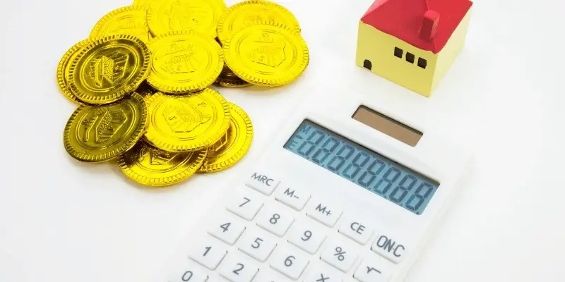 A white calculator, a small toy house with a red roof, and a pile of shiny gold coins arranged on a clean white surface.