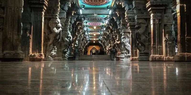 A low angle view of an ornate temple hallway with carved stone pillars and colorful lights on the ceiling reflecting on the polished floor.