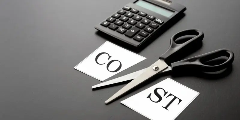 Scissors cutting a paper labeled “cost” beside a calculator on a dark surface.