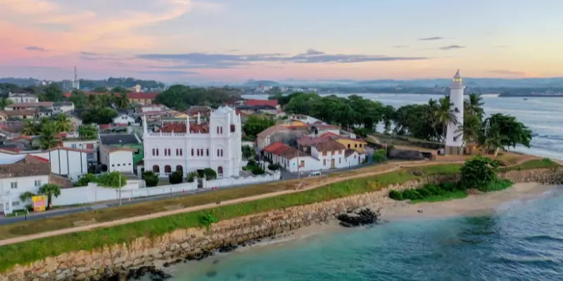 A seaside view showing historic buildings, palm trees, and gentle waves along a stone wall.