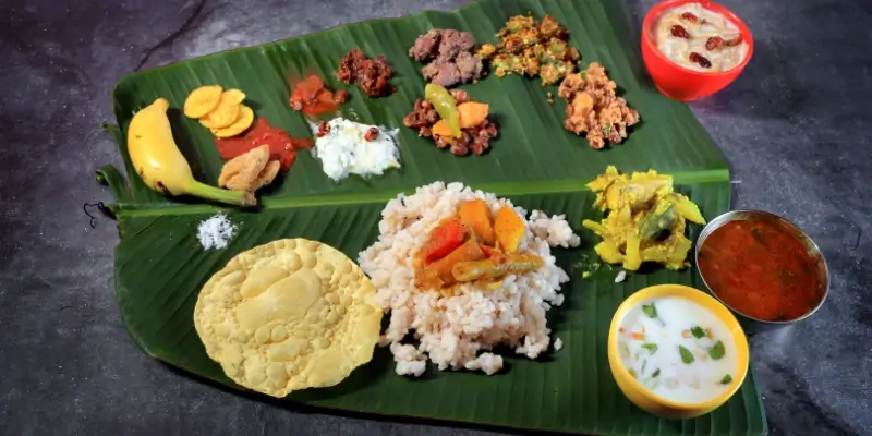 A traditional Indian meal arranged neatly on a large banana leaf, featuring rice, papad, banana, pickles, mixed vegetable dishes, curries, yogurt, and a sweet dessert.