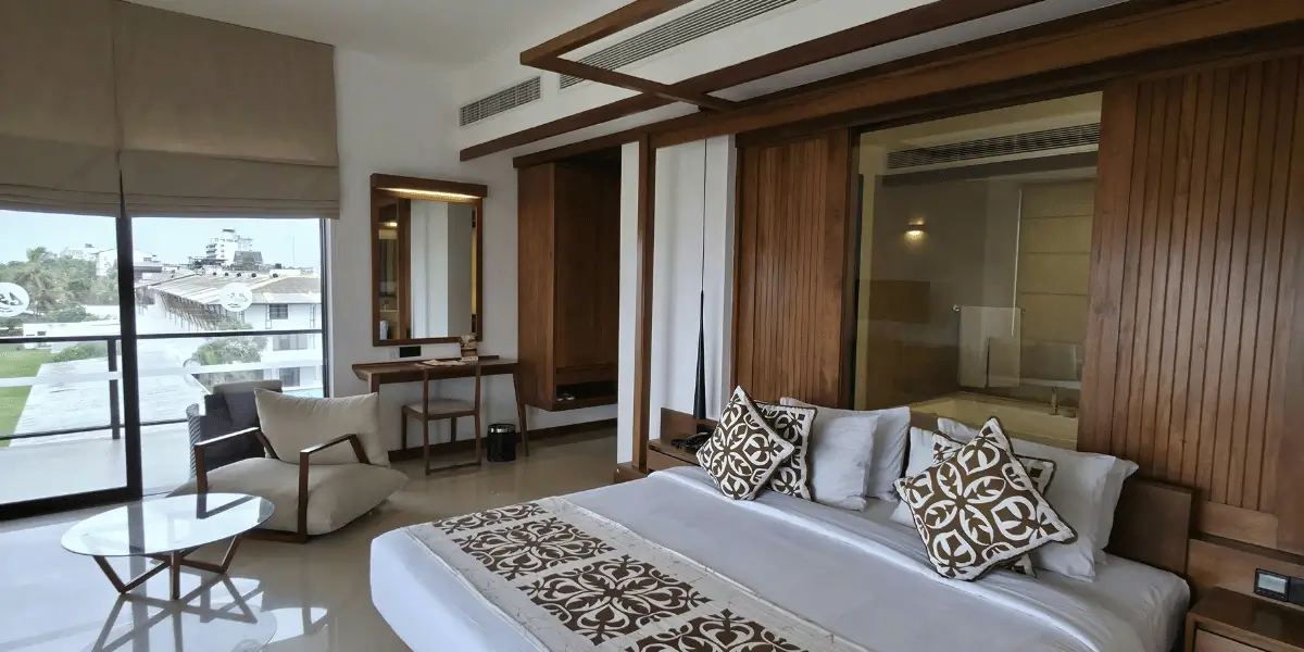 A bright hotel room with a large bed, patterned pillows, wooden walls, a glass partition, a cozy chair with a small round table, and a desk facing a wide window overlooking buildings and greenery.