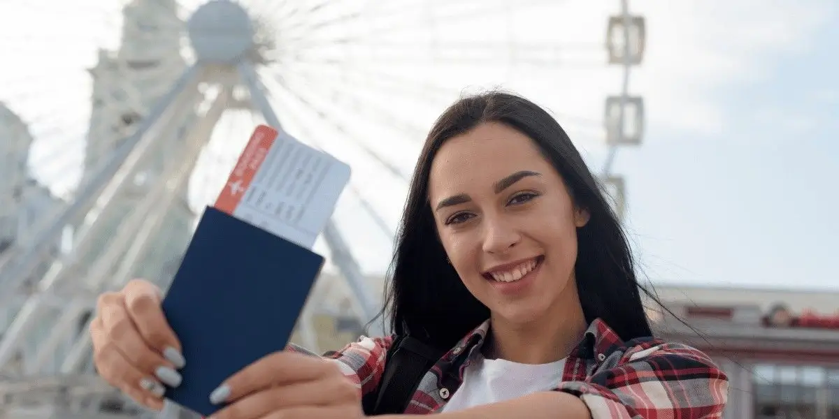 A young woman smiling and holding a passport with a boarding pass in front of a large Ferris wheel outdoors.