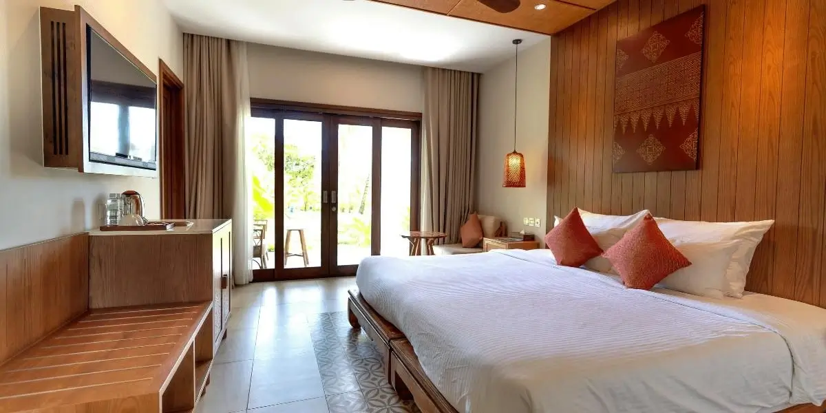 A cozy hotel room with a large bed, warm wooden walls, soft lighting, and glass doors opening to a bright outdoor view.