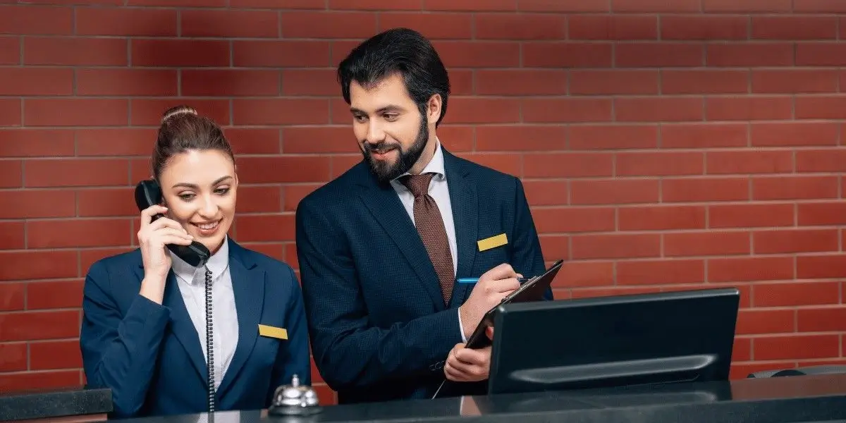 Two hotel reception staff standing at the front desk, one speaking on the phone and the other holding a clipboard.
